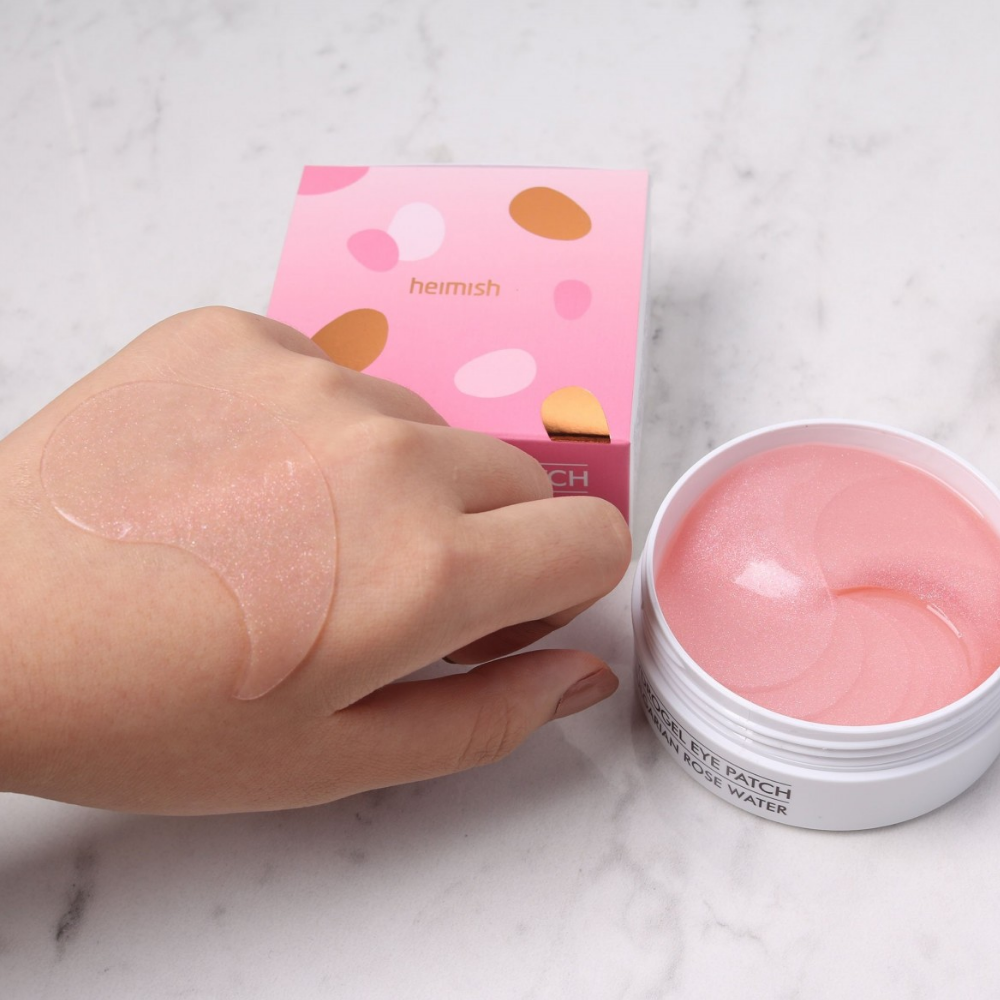 Eine Hand hält eine rosa Bulgarian Rose Hydrogel Eye Patch 60st von heimish und trägt eine Augenmaske auf.