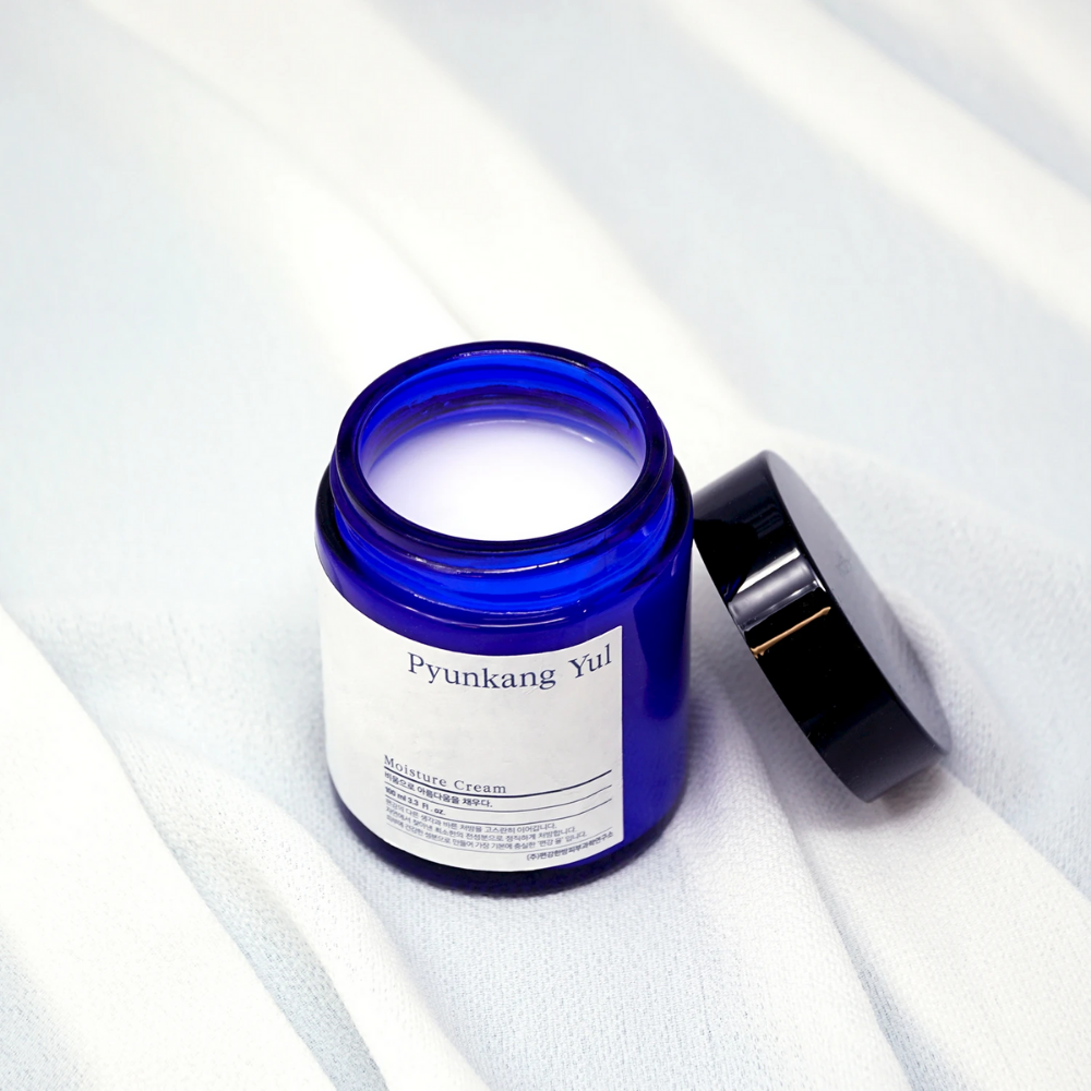 Eine blaue Dose mit Deckel, die auf einem weißen Tuch sitzt und die feuchtigkeitsspendenden Eigenschaften von Pyunkang Yul's Moisture Cream 100ml zeigt.