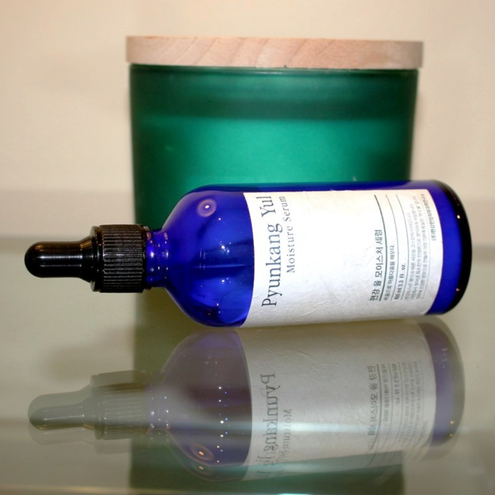 Eine Flasche Pyunkang Yul Moisture Serum 100ml gleicht die Feuchtigkeitsniveaus auf einem Tisch neben einer Kerze aus.