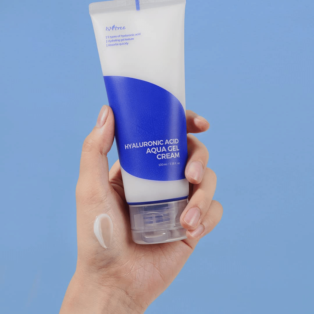 Eine Hand, die eine Tube Isntree Hyaluronic Acid Aqua Gel Cream 100ml vor einem blauen Hintergrund hält.